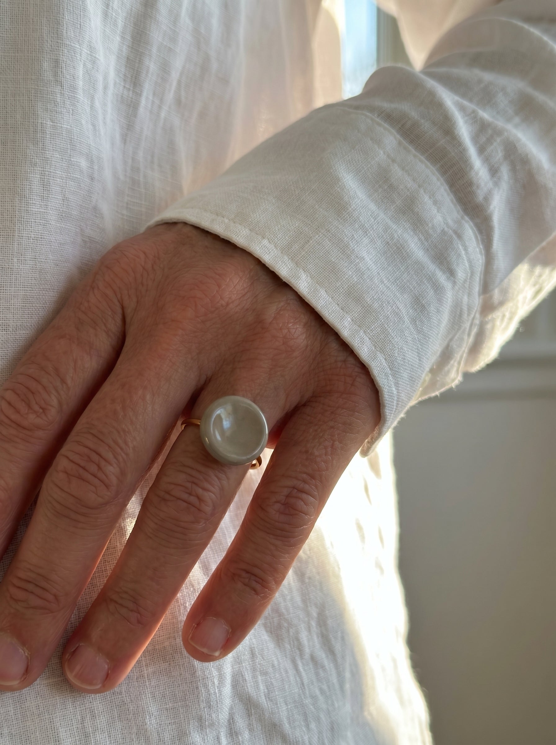 Nidus Topo — anillo de porcelana con cúpula circular en esmalte topo brillante en mano con camisa lino blanca, luz solar lateral suave