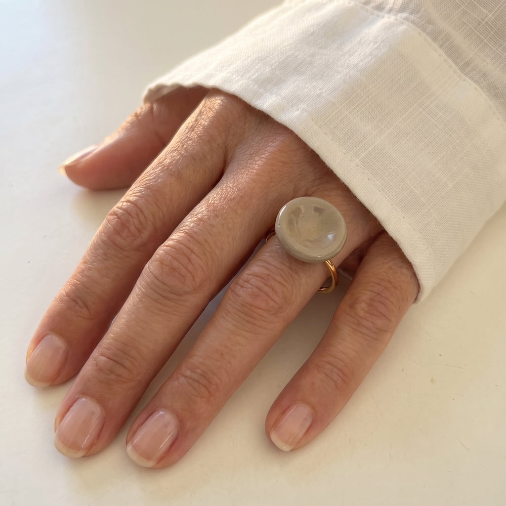 Nidus Topo — anillo de porcelana con cúpula circular en esmalte topo brillante en mano apoyada sobre superficie blanca con camisa lino, luz natural