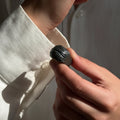 Onda Basalto — anillo de porcelana negra brillante con estrías verticales talladas a mano sostenido entre dedos con camisa blanca, fondo gris con luz lateral