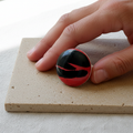 Magma — anillo de porcelana negro con vetas rojo carmín sobre baldosa de piedra beige, primer plano