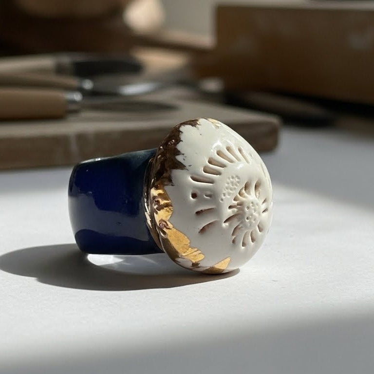 Caracola sobre el Mar — anillo de porcelana calada blanca con detalles dorados sobre banda azul marino sostenido entre dedos sobre superficie de piedra