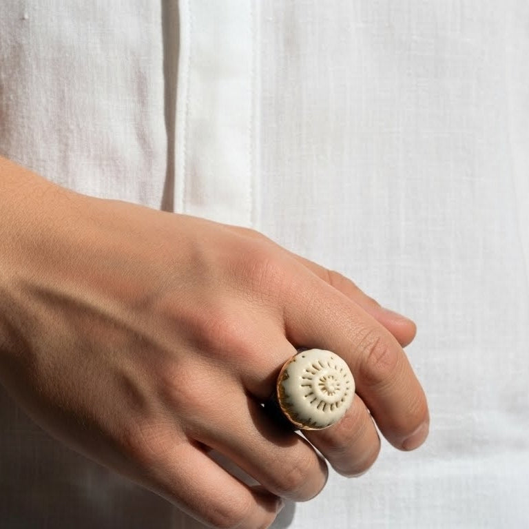 Caracola sobre el Mar — anillo de porcelana calada blanca con detalles dorados sobre banda azul marino sobre superficie blanca con luz solar lateral