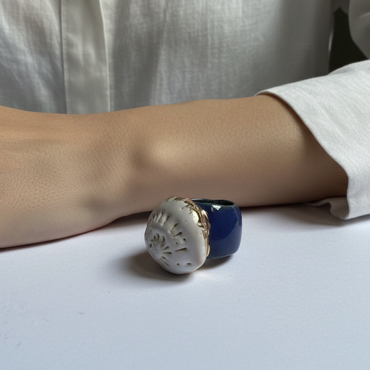 Caracola sobre el Mar — anillo de porcelana calada blanca con detalles dorados sobre banda azul marino sobre superficie blanca con sombra solar, vista frontal