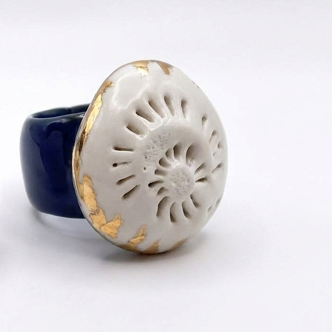 Caracola sobre el Mar — anillo de porcelana calada blanca con detalles dorados sobre banda azul marino apoyado sobre brazo con camisa blanca, vista cenital