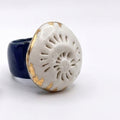 Caracola sobre el Mar — anillo de porcelana calada blanca con detalles dorados sobre banda azul marino apoyado sobre brazo con camisa blanca, vista cenital