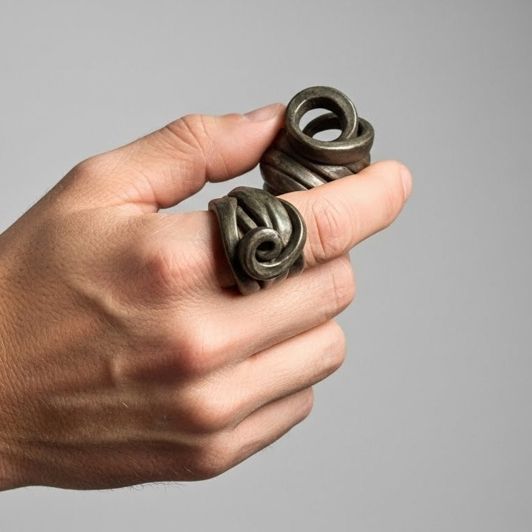 Peine Bronce — dos anillos de barro negro entrelazados en mano cerrada sobre fondo gris, vista frontal mostrando volumen escultórico