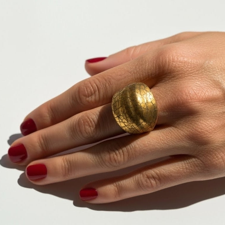 Crépité — anillo de barro terracota con cúpula de oro craquelado en mano con uñas rojas sobre suelo blanco con luz solar, vista cenital