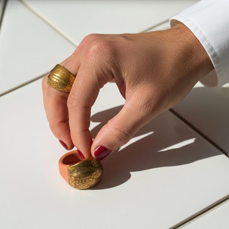 Crépité — anillo de barro terracota con cúpula de oro craquelado en mano con uñas rojas, vista lateral con sombra solar sobre suelo blanco