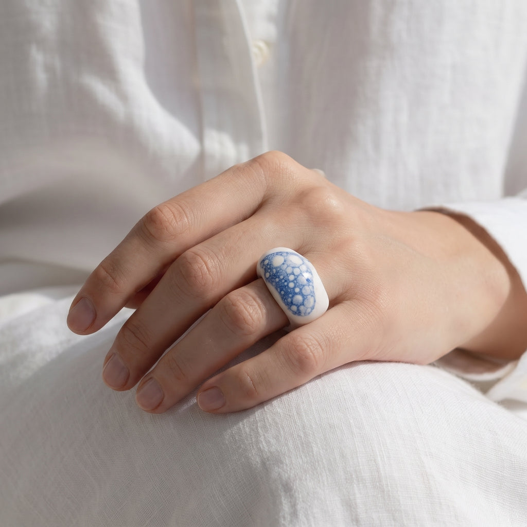 Espuma de Mar — anillo de porcelana blanca con esmalte azul y burbujas en mano apoyada sobre tela lino blanca, vista cenital con luz suave