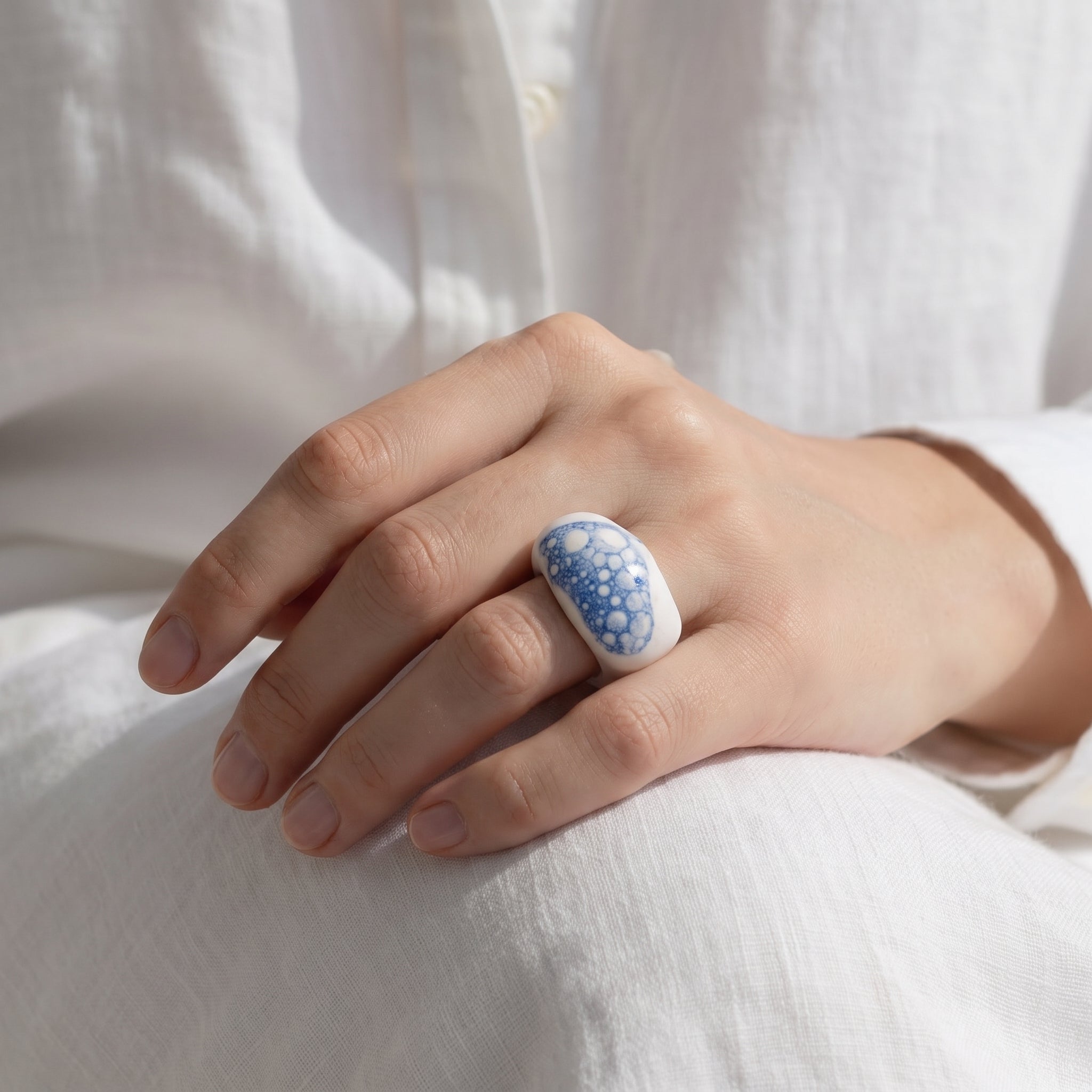 Espuma de Mar — anillo de porcelana blanca con esmalte azul y burbujas en mano apoyada sobre tela lino blanca, vista cenital con luz suave