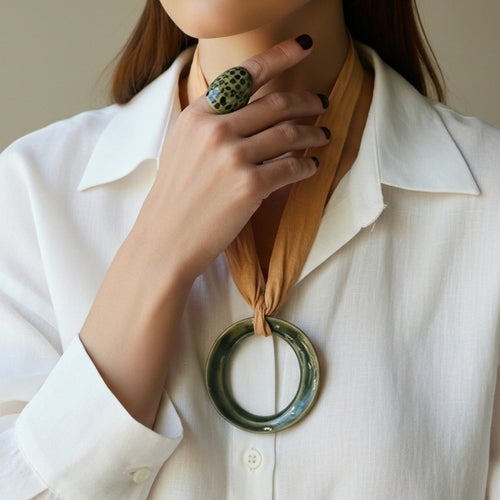 Áspid Anillo — anillo de porcelana verde musgo con manchas turquesa en mano apoyada en cuello con camisa blanca, colgante Áspid visible al fondo