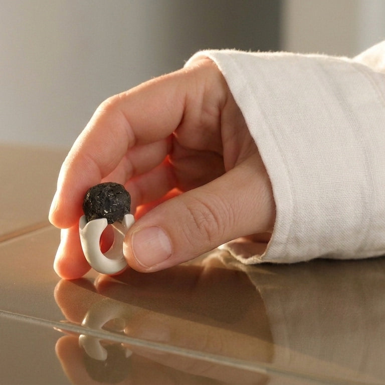 Antithétikós — anillo de porcelana blanca con masa negra rugosa apoyado sobre superficie reflectante con camisa blanca, luz cálida lateral