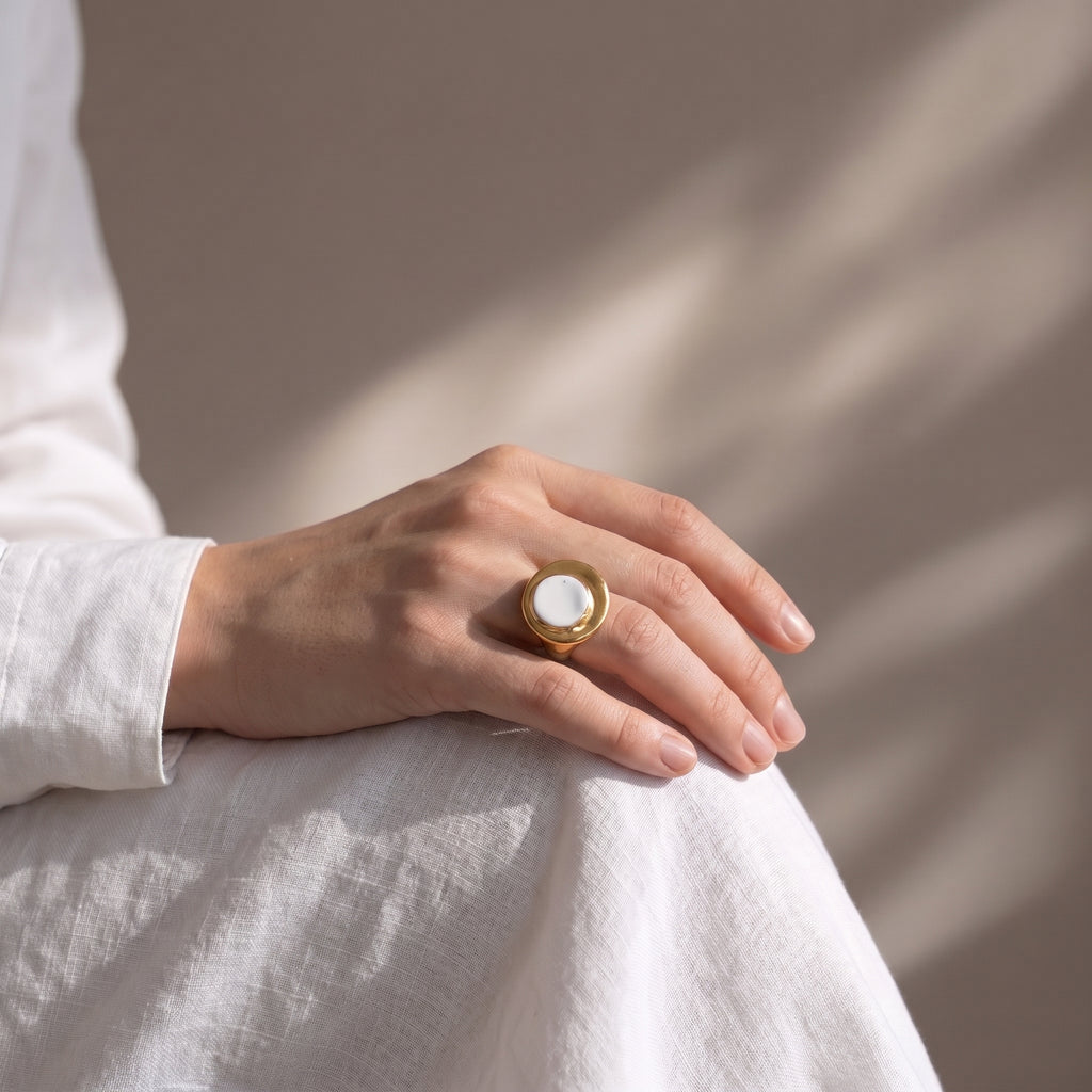 Eclipse Parcial — anillo de porcelana blanca con aro de oro ajustable en mano apoyada sobre tela lino blanca, luz de ventana con sombras de hojas sobre fondo beige