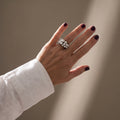 Burbujas Black — anillo de porcelana negra con esferas blancas apiladas en mano con uñas burdeos y camisa blanca, fondo beige con luz lateral