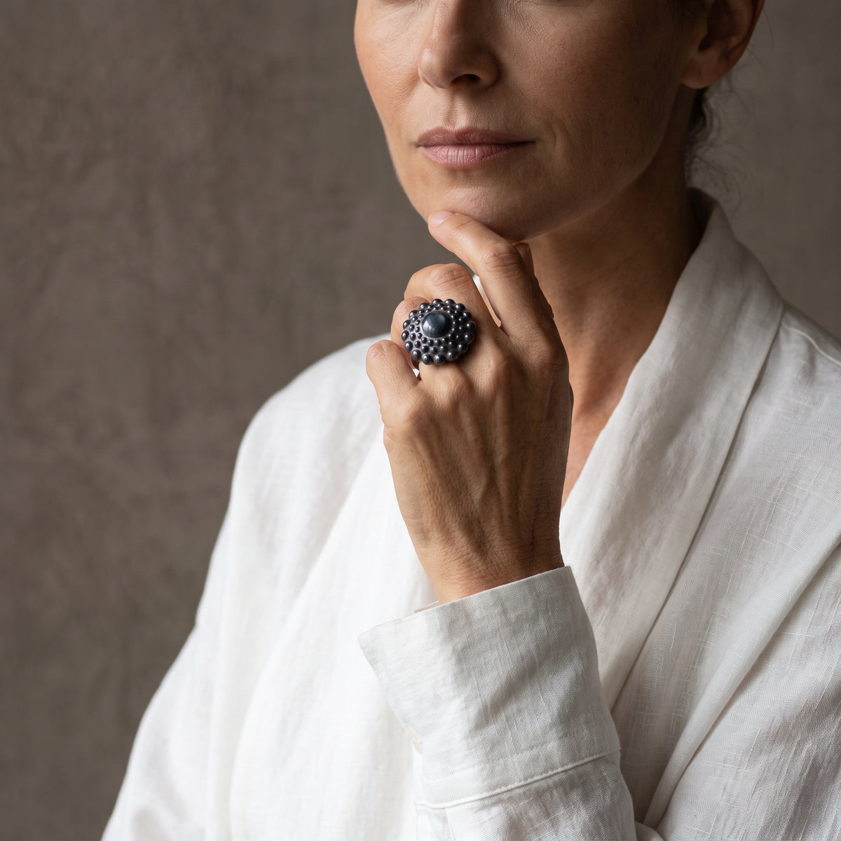 Girándola Azul Nacarado — mujer con camisa blanca sosteniendo su barbilla con la mano, anillo de porcelana azul pizarra nacarado en el índice, fondo gris cálido