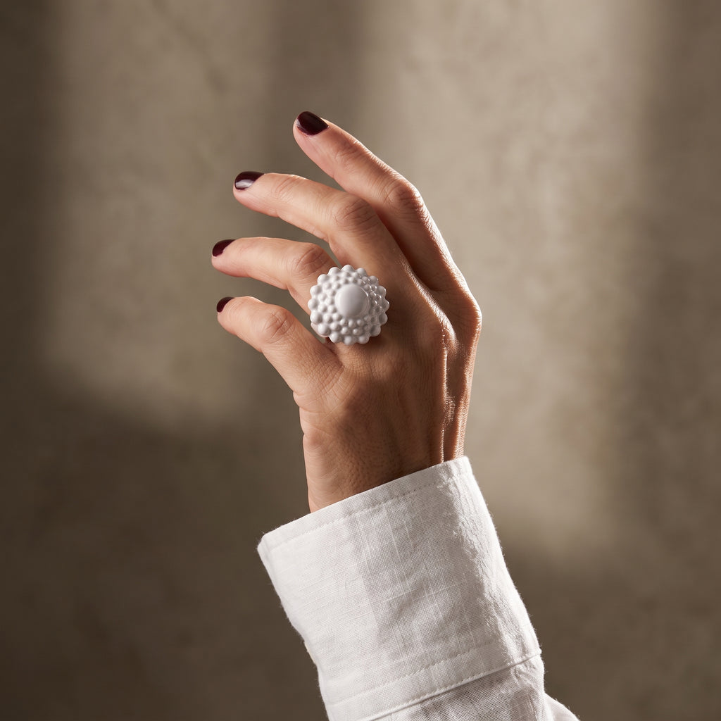 Girándola Blanco Nacarado — mano con uñas granate y camisa blanca mostrando el anillo de porcelana blanca nacarada en el índice, fondo gris cálido