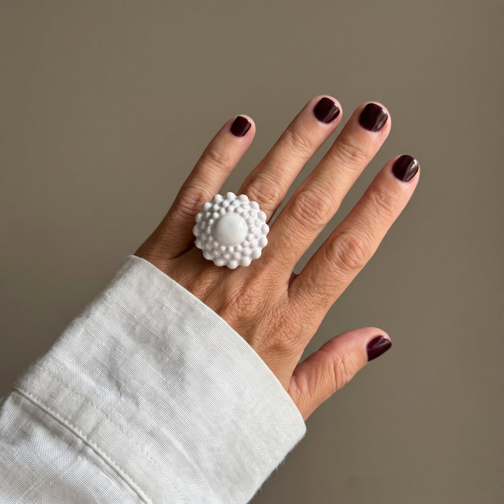 Girándola Blanco Nacarado — mano elevada con camisa blanca y anillo de porcelana blanca nacarada, fondo beige con luz suave
