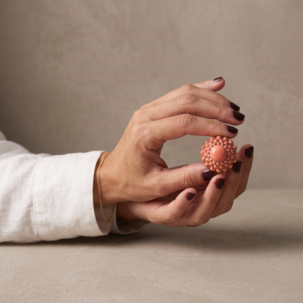 Girándola Coral — dos manos sosteniendo el anillo de porcelana coral con bultos dorados, camisa blanca de lino, fondo beige cálido