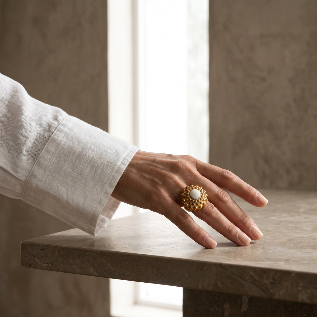 Girándola Amanecer — mano femenina con camisa blanca apoyada sobre superficie de mármol beige, anillo dorado rosado con centro blanco, luz natural
