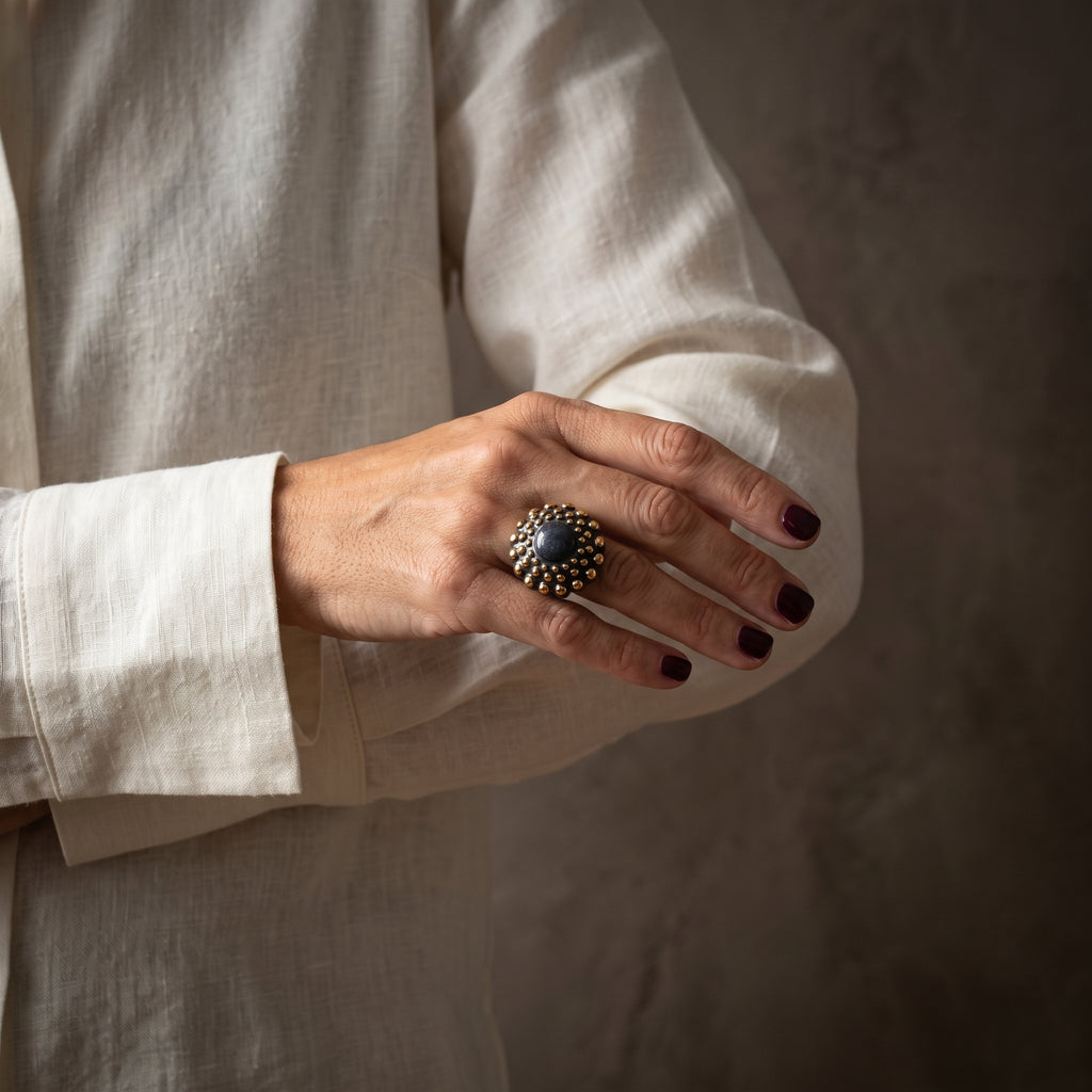 Girándola Pizarra Oro — mano con uñas granate y camisa blanca mostrando el anillo pizarra dorado en el índice, fondo beige cálido