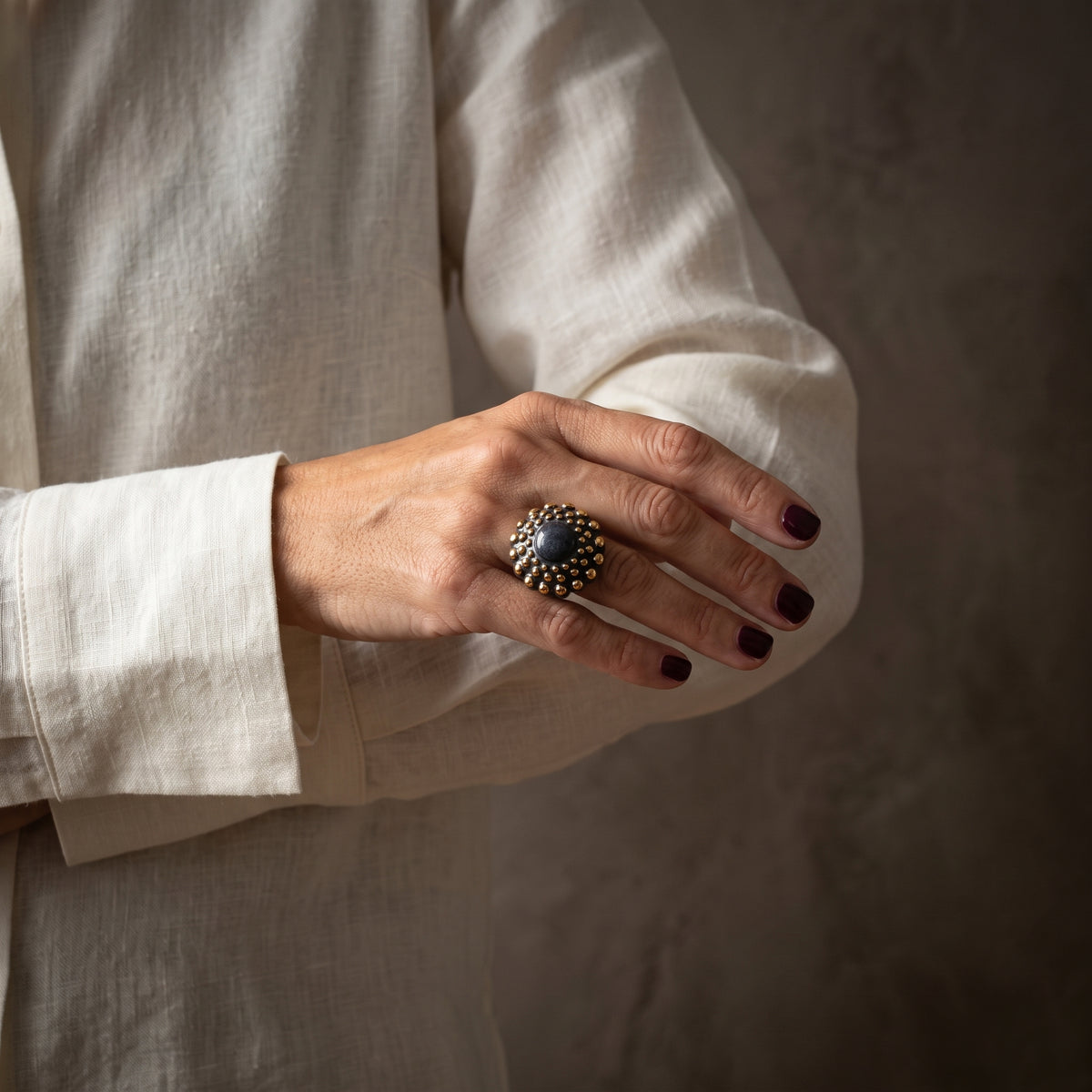 Girándola Pizarra Oro — mano con uñas granate y camisa blanca mostrando el anillo pizarra dorado en el índice, fondo beige cálido