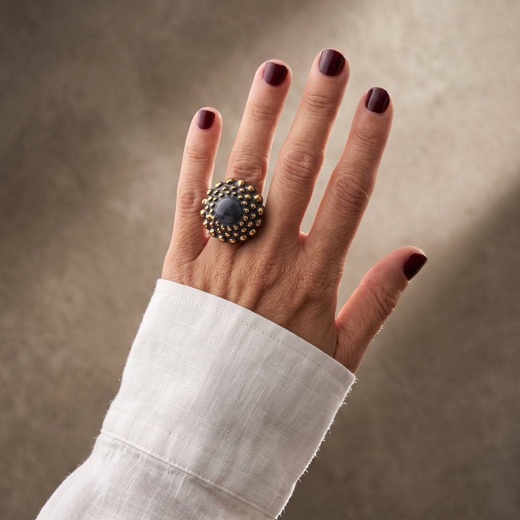 Girándola Pizarra Oro — mano con camisa blanca de lino cruzada, anillo de porcelana pizarra con bultos dorados, fondo beige texturizado