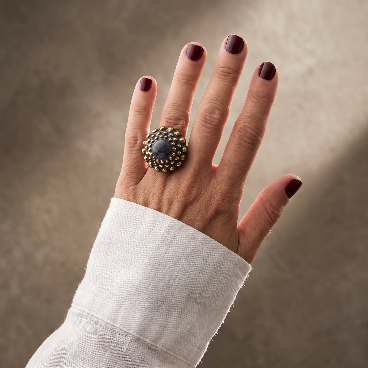 Girándola Pizarra Oro — mano con camisa blanca de lino cruzada, anillo de porcelana pizarra con bultos dorados, fondo beige texturizado