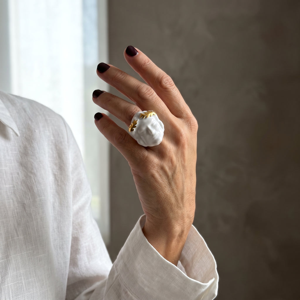 Dominus — mano femenina con camisa blanca alzada junto a ventana, anillo escultórico de porcelana blanca con corona de hojas doradas, luz natural lateral