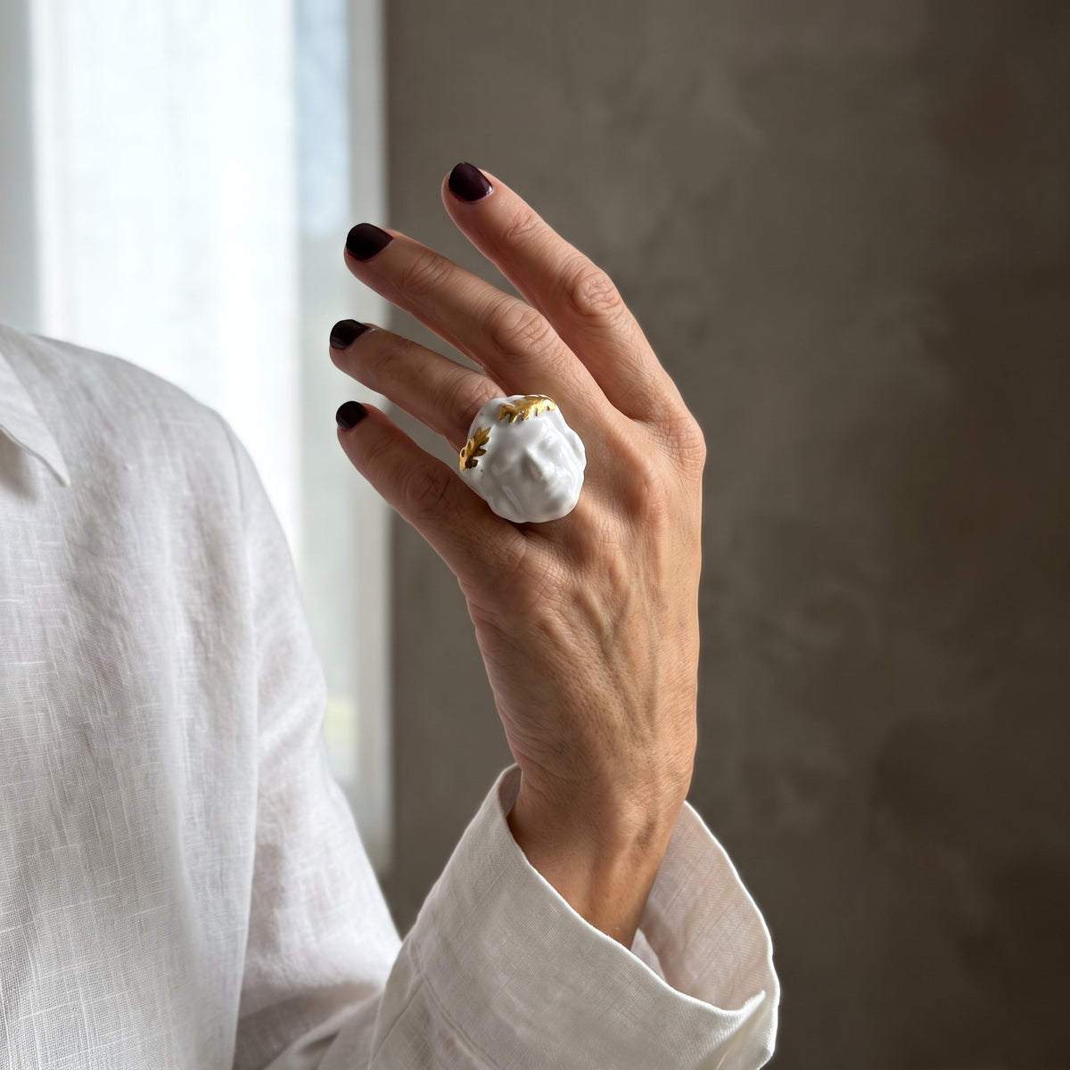 Dominus — mano femenina con camisa blanca alzada junto a ventana, anillo escultórico de porcelana blanca con corona de hojas doradas, luz natural lateral