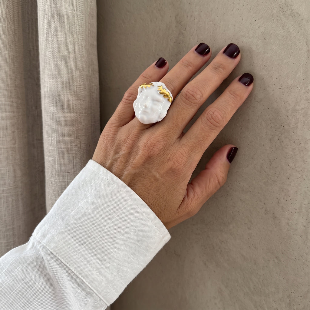 Dominus — mano femenina con camisa blanca apoyada sobre pared beige, anillo de porcelana con rostro masculino y guirnalda dorada en primer plano