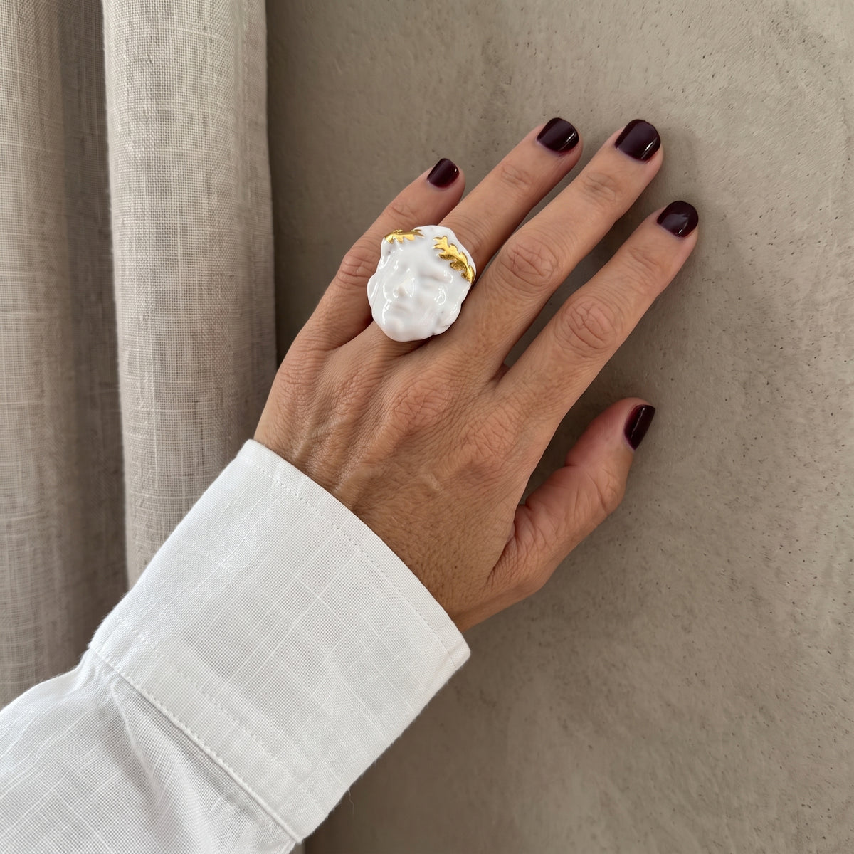 Dominus — mano femenina con camisa blanca apoyada sobre pared beige, anillo de porcelana con rostro masculino y guirnalda dorada en primer plano