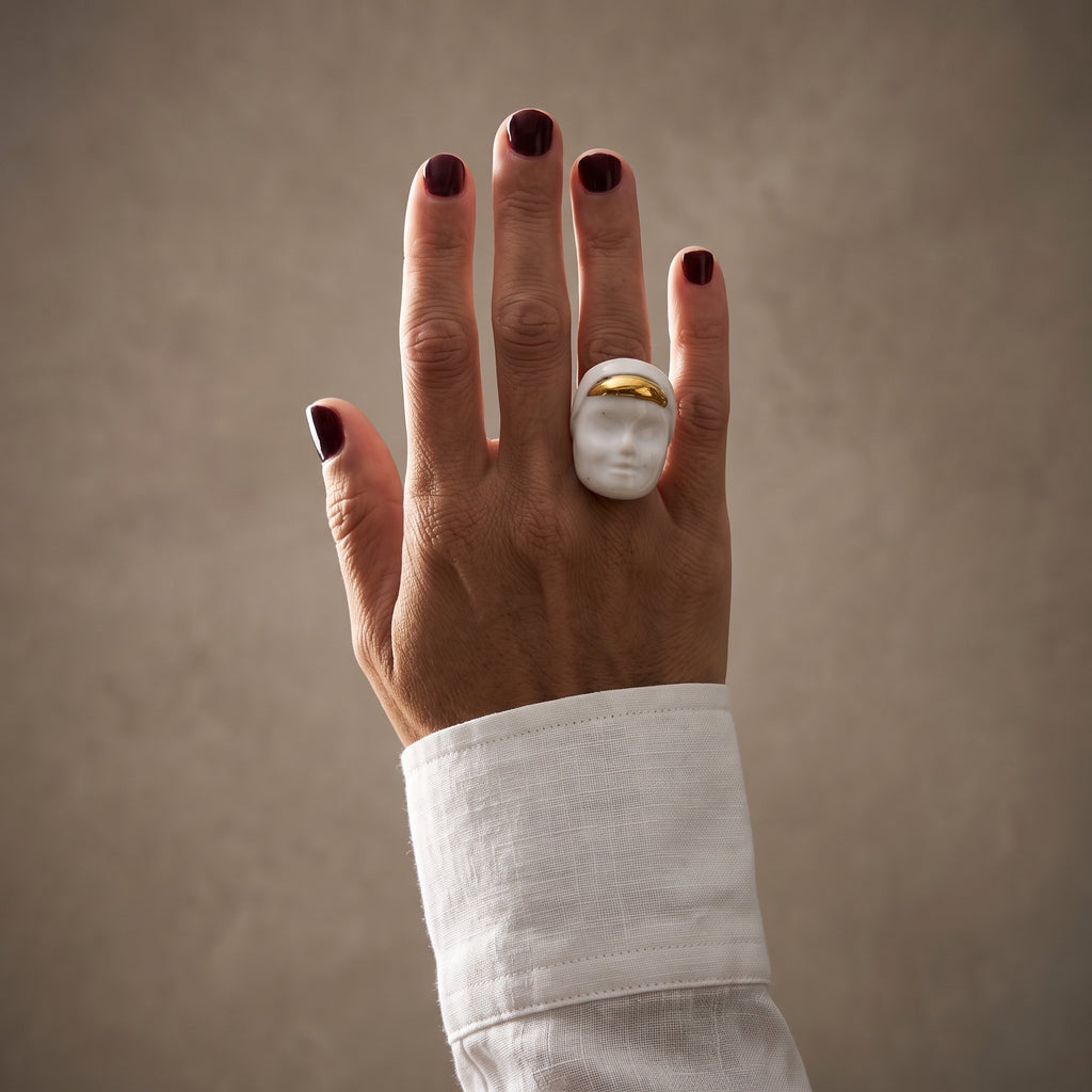 Domina — mano femenina con camisa blanca apoyada sobre hombro, anillo escultórico de porcelana blanca con franja dorada, luz natural