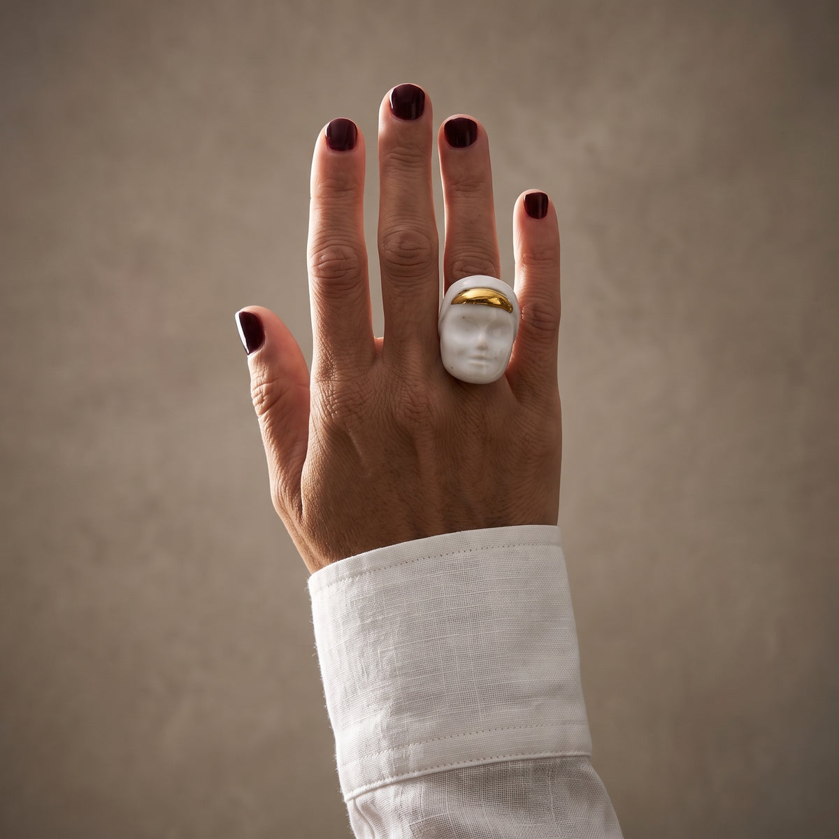 Domina — mano femenina con camisa blanca apoyada sobre hombro, anillo escultórico de porcelana blanca con franja dorada, luz natural