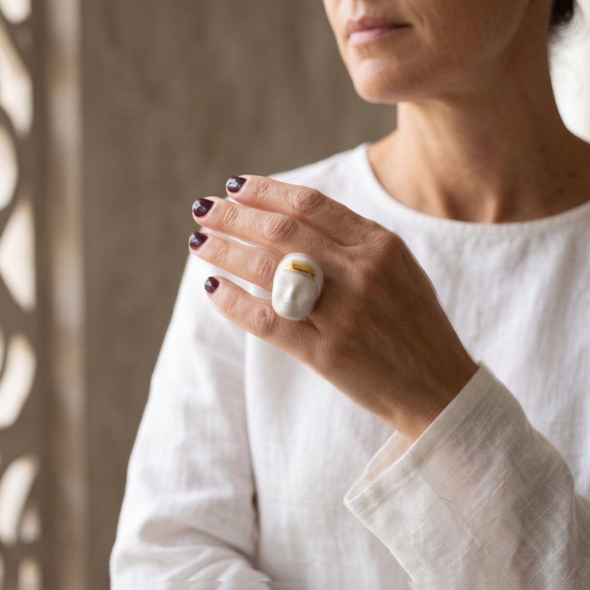 Domina — mano femenina con camisa blanca de lino alzada, anillo de porcelana con rostro y acabado dorado, fondo beige texturizado