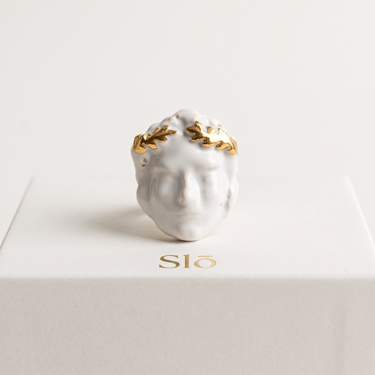 Dominus — vista frontal del anillo de porcelana con rostro y laurel dorado, presentado sobre caja de regalo Slō, fondo crema