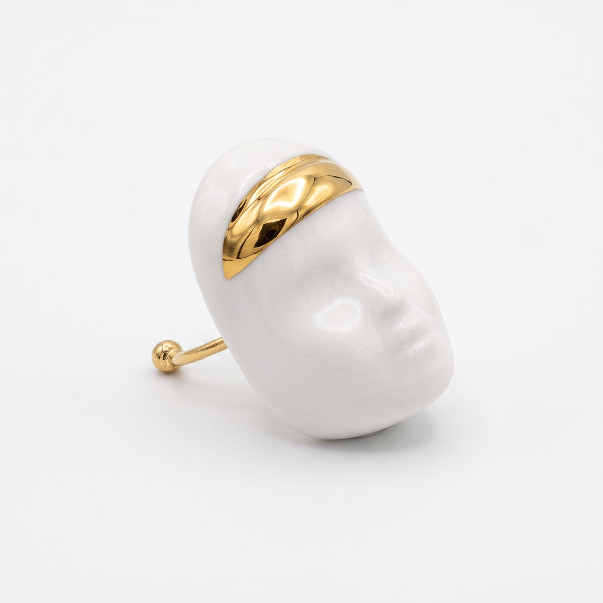 Domina — vista lateral del anillo escultórico de porcelana blanca con rostro y franja dorada en la frente, aro dorado abierto, fondo blanco