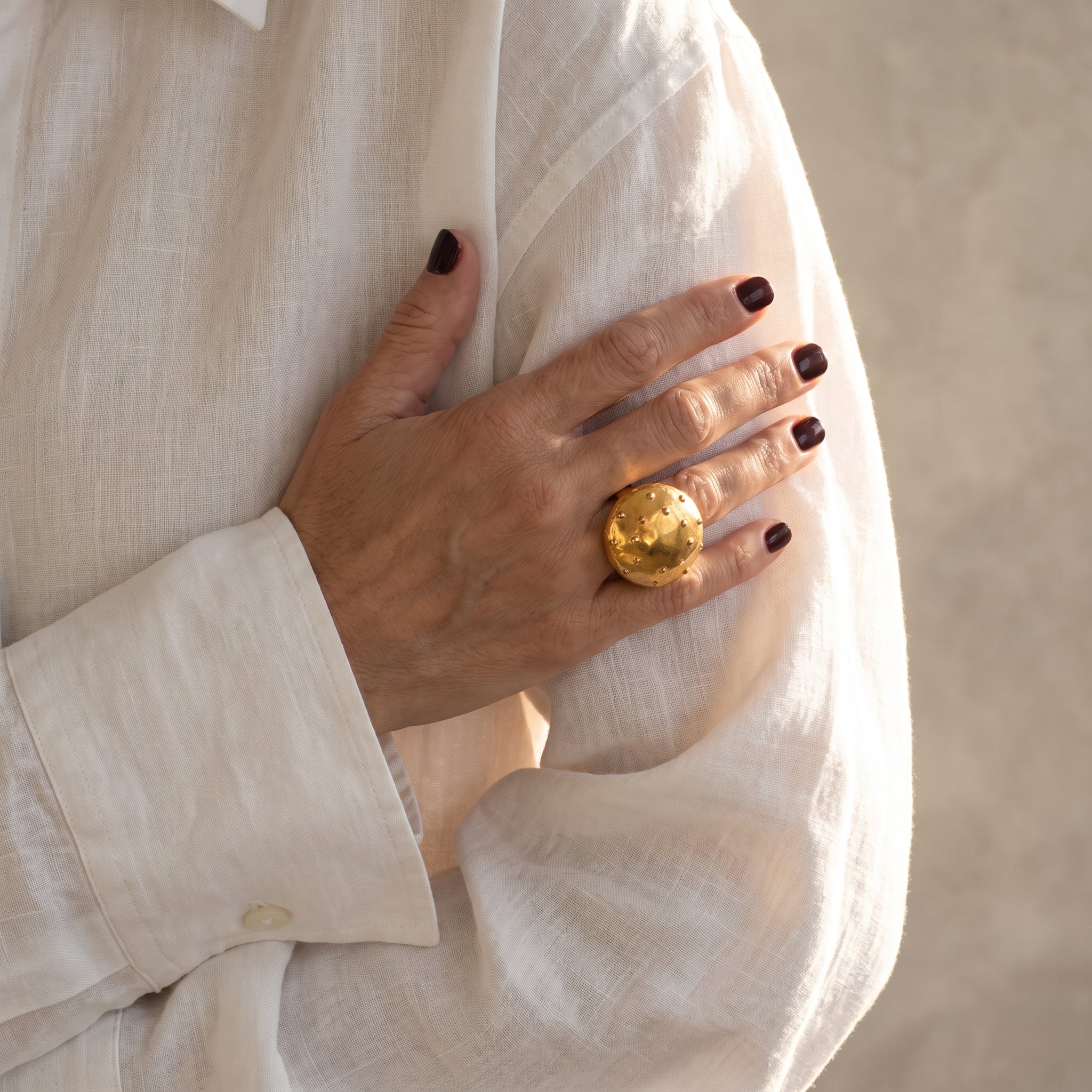 Erizo Dorado — anillo icónico de porcelana con cúpula dorada y púas esféricas en mano con uñas burdeos y camisa lino blanca, fondo beige con luz solar lateral