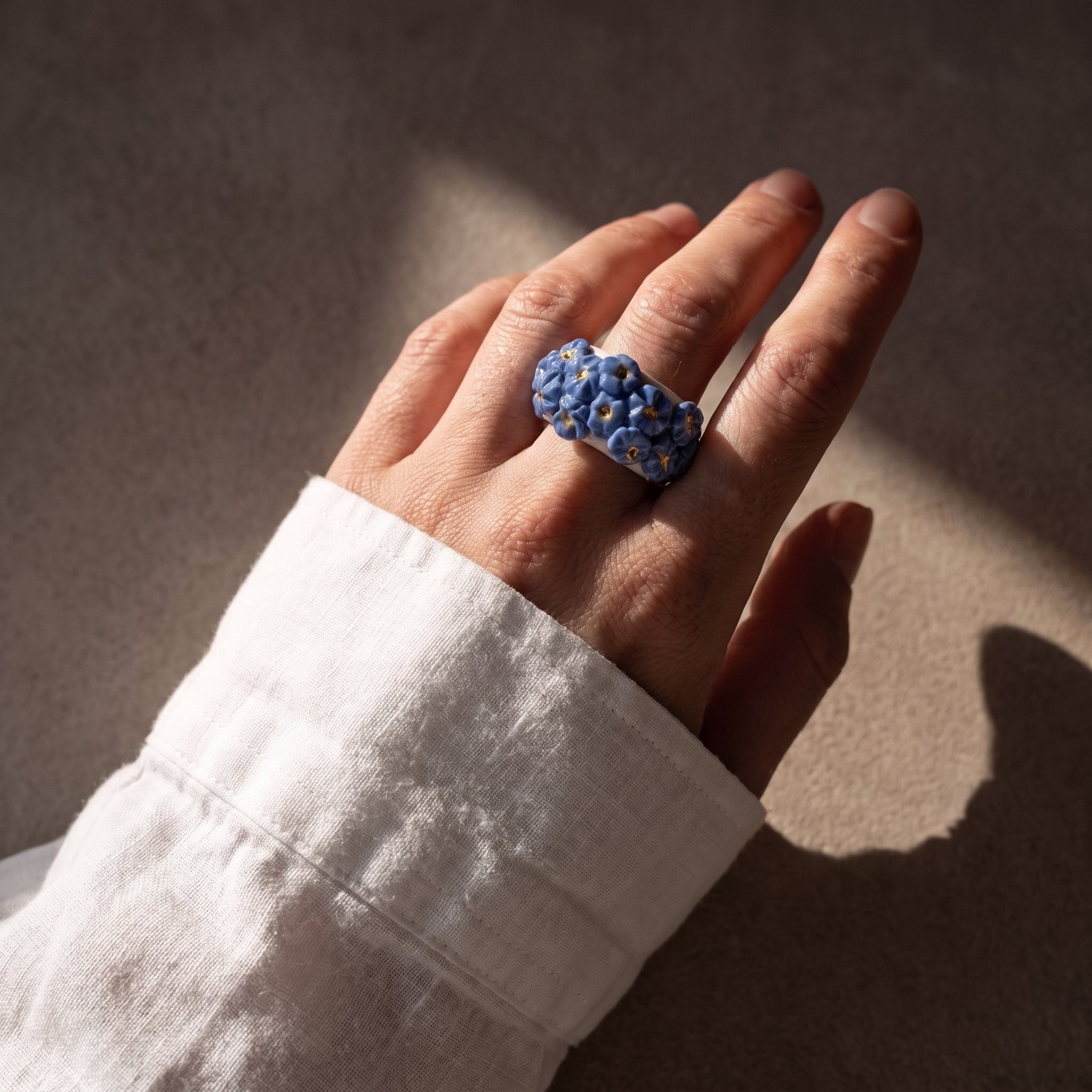 Bouquet Azul — anillo de porcelana con flores azul aciano en mano con camisa lino blanca, luz solar lateral sobre fondo tierra