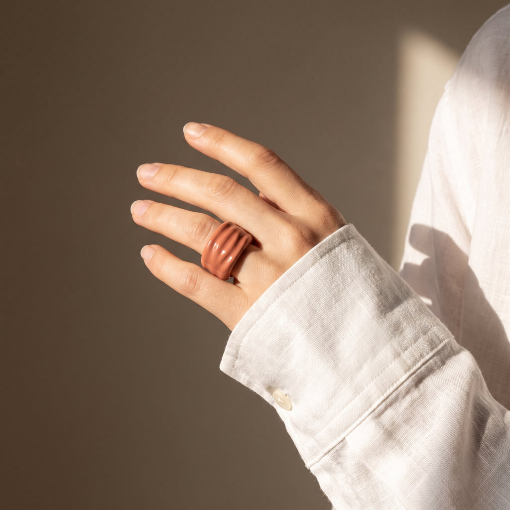 Onda Maquillaje — anillo de porcelana con esmalte terracota rosado brillante y estrías horizontales en mano alzada con camisa lino blanca, luz solar cálida con sombras sobre fondo tierra
