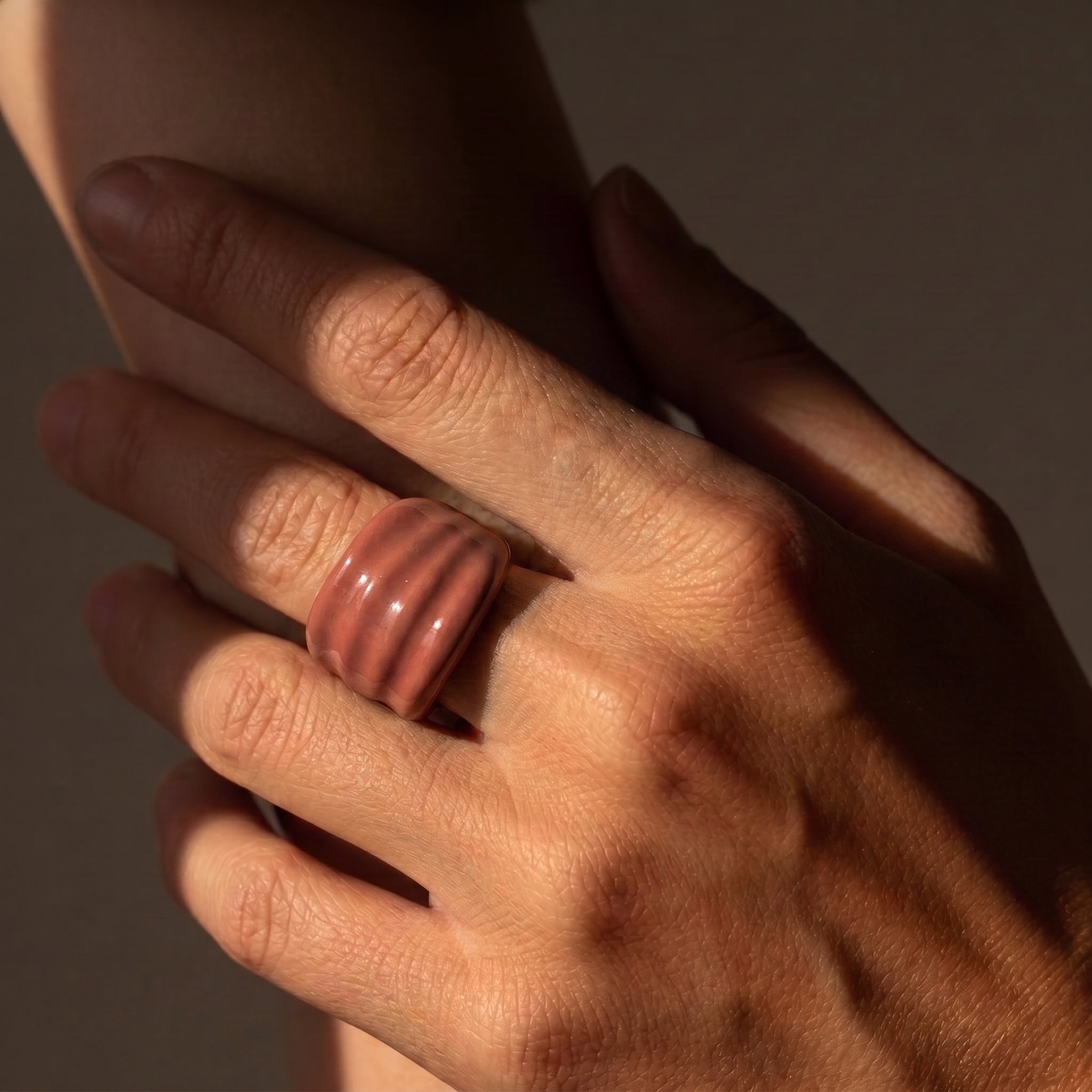 Onda Maquillaje — anillo de porcelana con esmalte terracota rosado brillante y estrías horizontales en mano apoyada sobre otra mano, luz solar lateral con sombras dramáticas sobre fondo oscuro