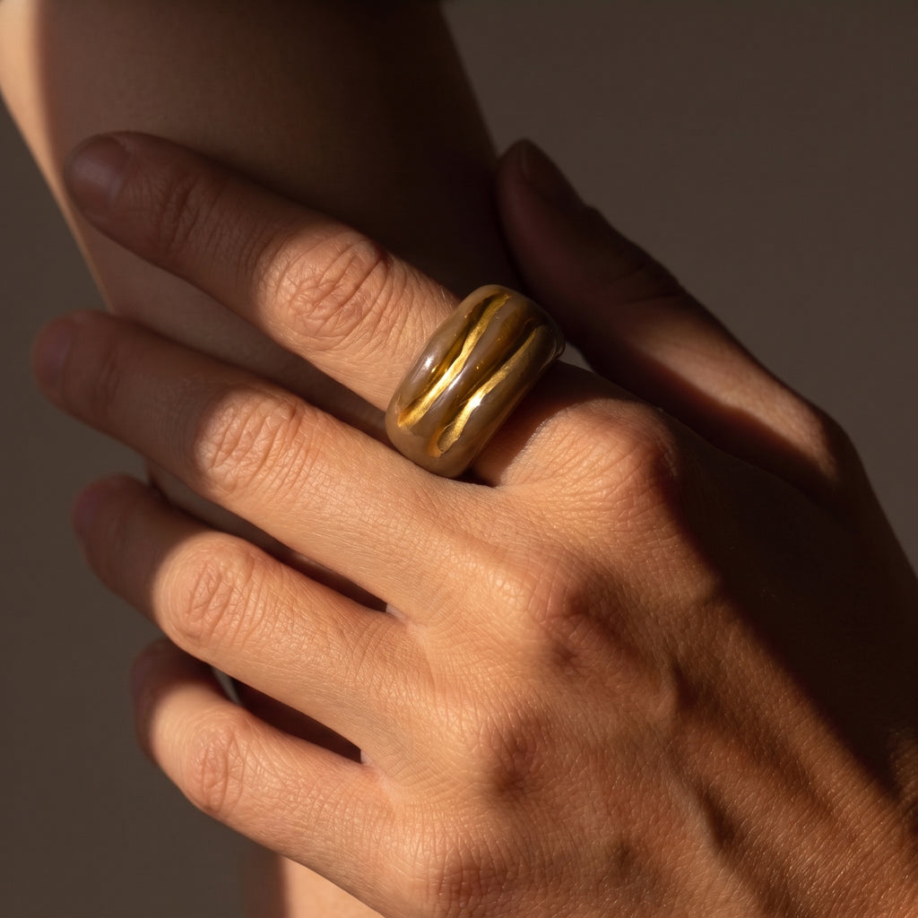 Onda Caramelo — anillo de porcelana con esmalte caramelo brillante y vetas doradas en mano con uñas burdeos y camisa lino blanca, fondo beige con luz solar lateral