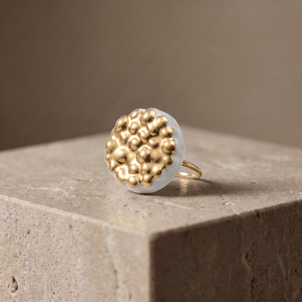 Maní Dorado — anillo de porcelana blanca con cúpula circular cubierta de esferas doradas sobre aro fino dorado, sobre superficie de piedra caliza con fondo neutro beige