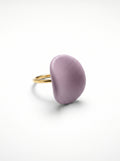 Pétalo Morado — anillo ajustable de porcelana lavanda mate con aro de oro en mano apoyada sobre superficie beige, vista cenital con luz natural