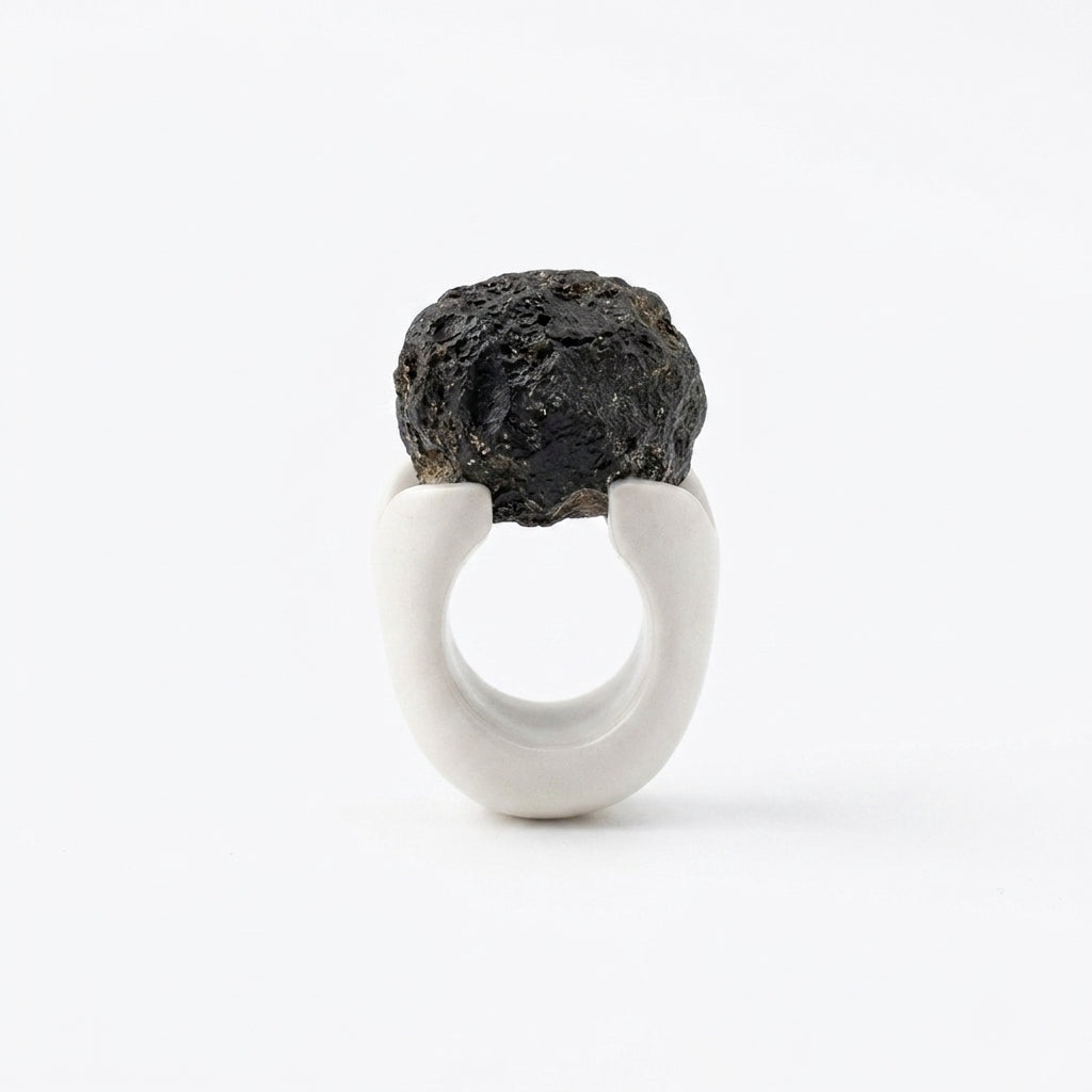 Antithétikós — anillo de porcelana blanca mate con masa negra rugosa volcánica encima, vista frontal sobre fondo blanco mostrando el contraste total entre texturas