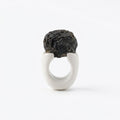 Antithétikós — anillo de porcelana blanca mate con masa negra rugosa volcánica encima, vista frontal sobre fondo blanco mostrando el contraste total entre texturas