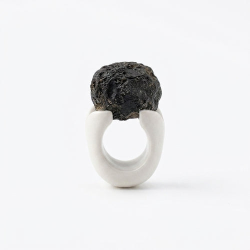 Antithétikós — anillo de porcelana blanca mate con masa negra rugosa volcánica encima, vista frontal sobre fondo blanco mostrando el contraste total entre texturas