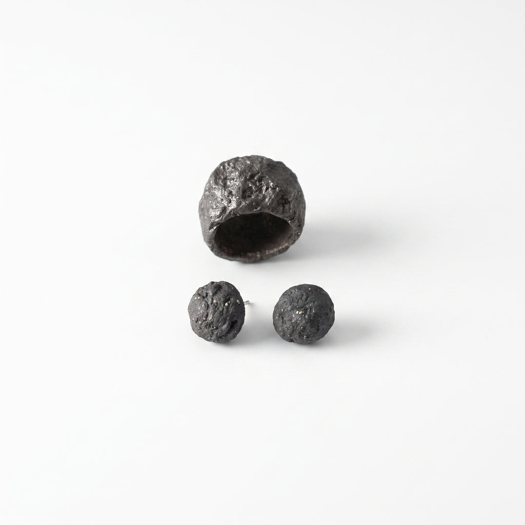 Grava Puzolana Negra — anillo y pendientes de barro negro de alta temperatura sobre fondo blanco, vista cenital con textura volcánica detallada