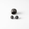Grava Puzolana Negra — anillo y pendientes de barro negro de alta temperatura sobre fondo blanco, vista cenital con textura volcánica detallada