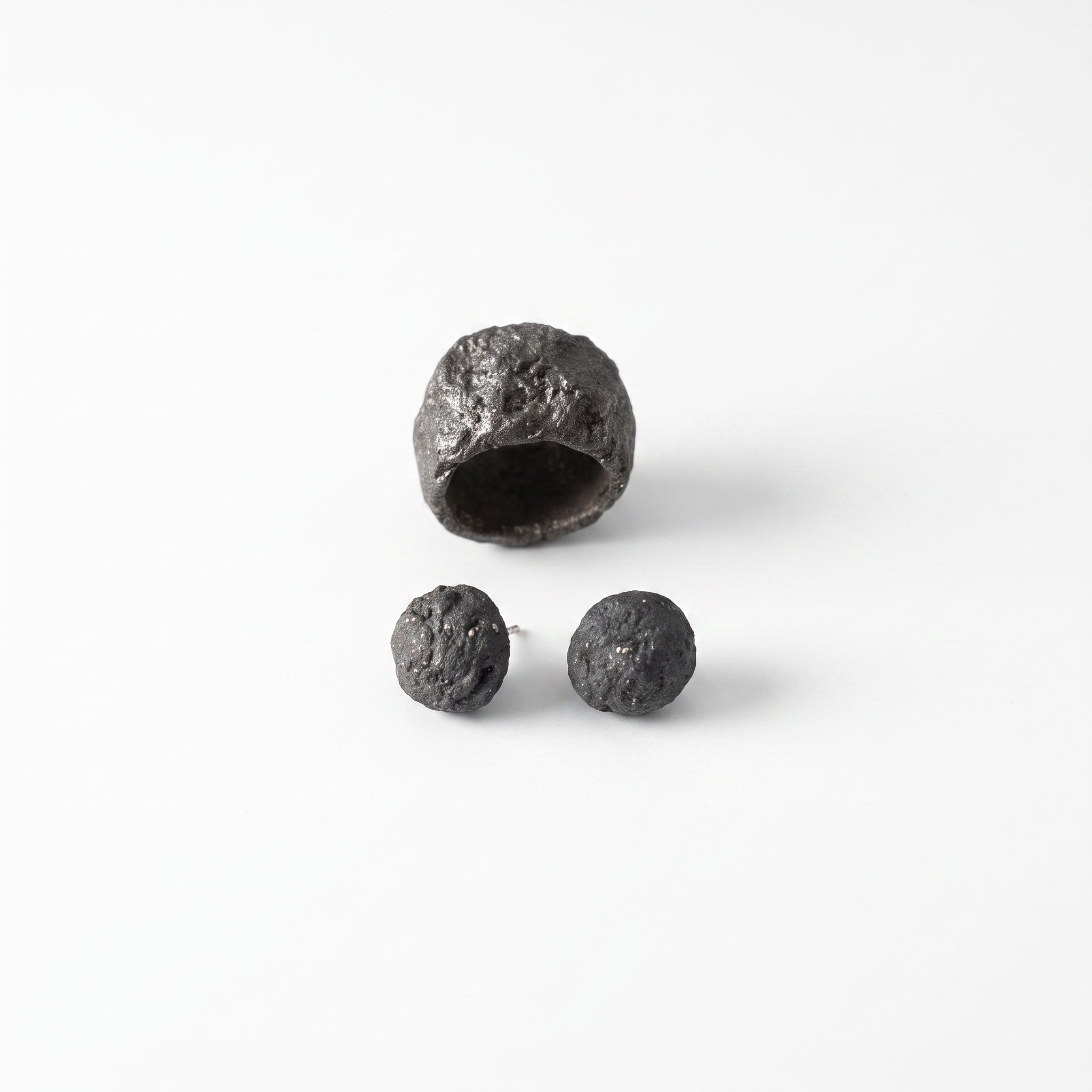 Grava Puzolana Negra — anillo y pendientes de barro negro de alta temperatura sobre fondo blanco, vista cenital con textura volcánica detallada