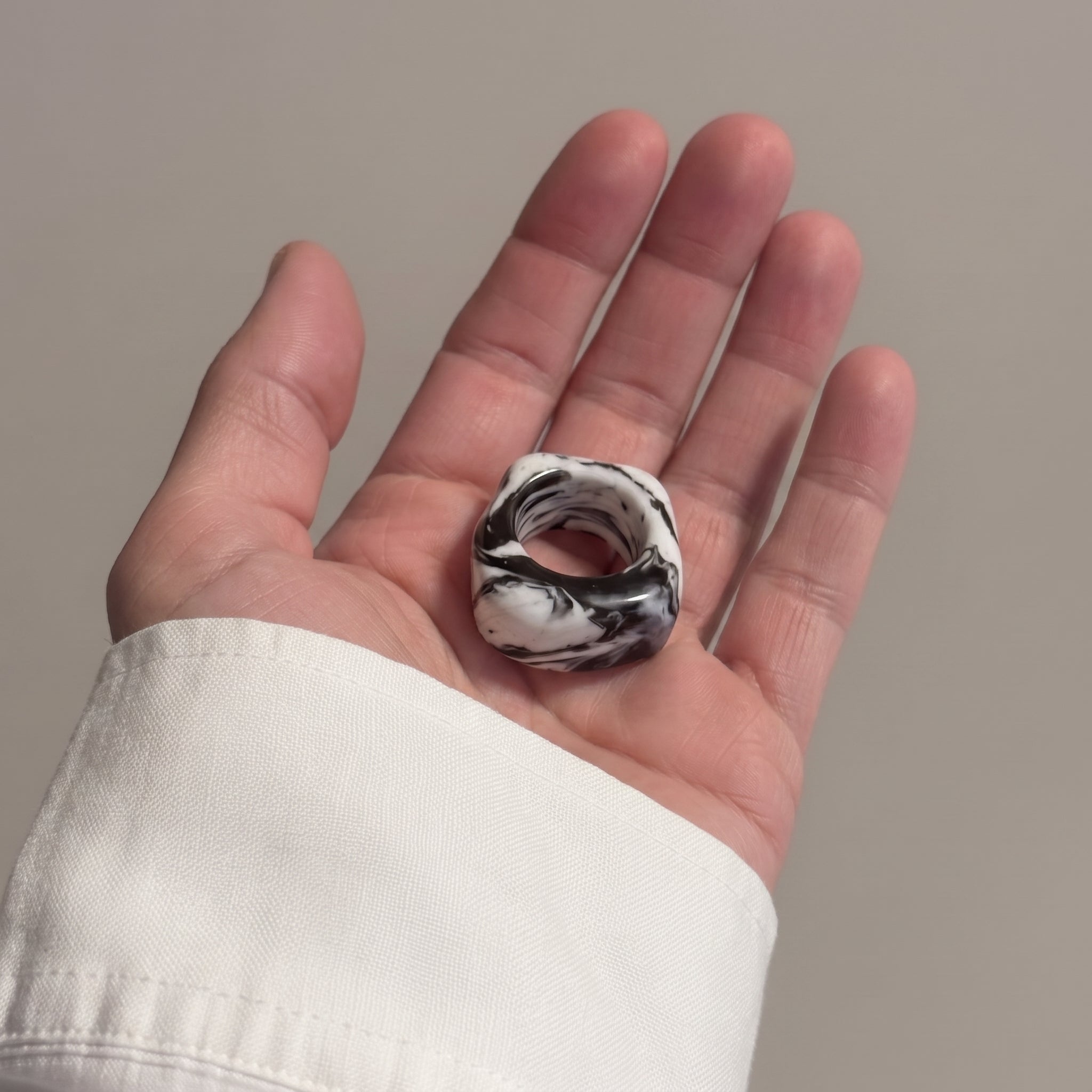 Petra Surco Black — anillo de porcelana negro con vetas blancas marmoladas en palma de mano abierta con camisa blanca, fondo gris beige